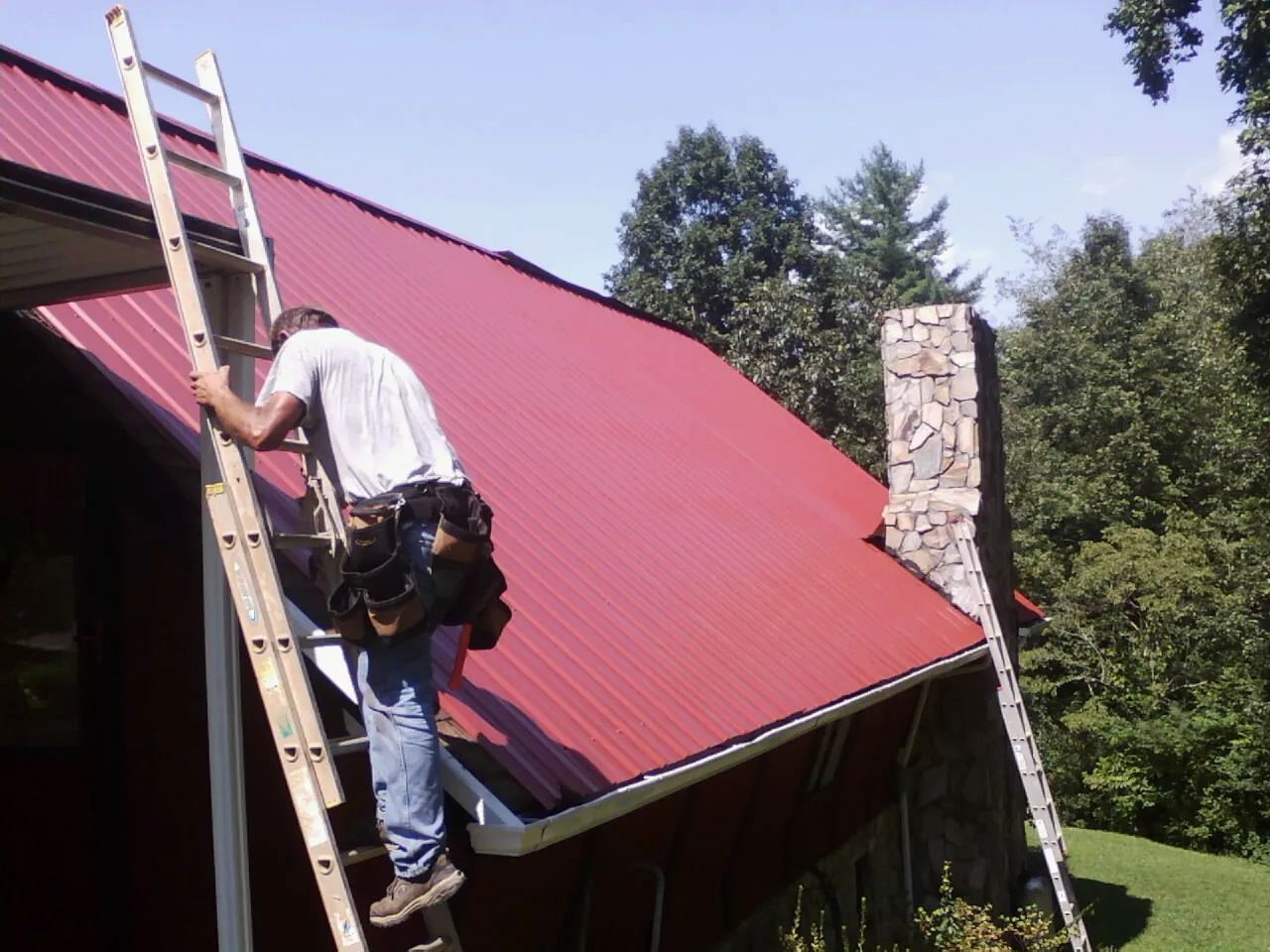 Roofer on a ladder beside a red metal roof and stone chimney — Soffit & Fascia Repair by Roofing Brothers in Albemarle